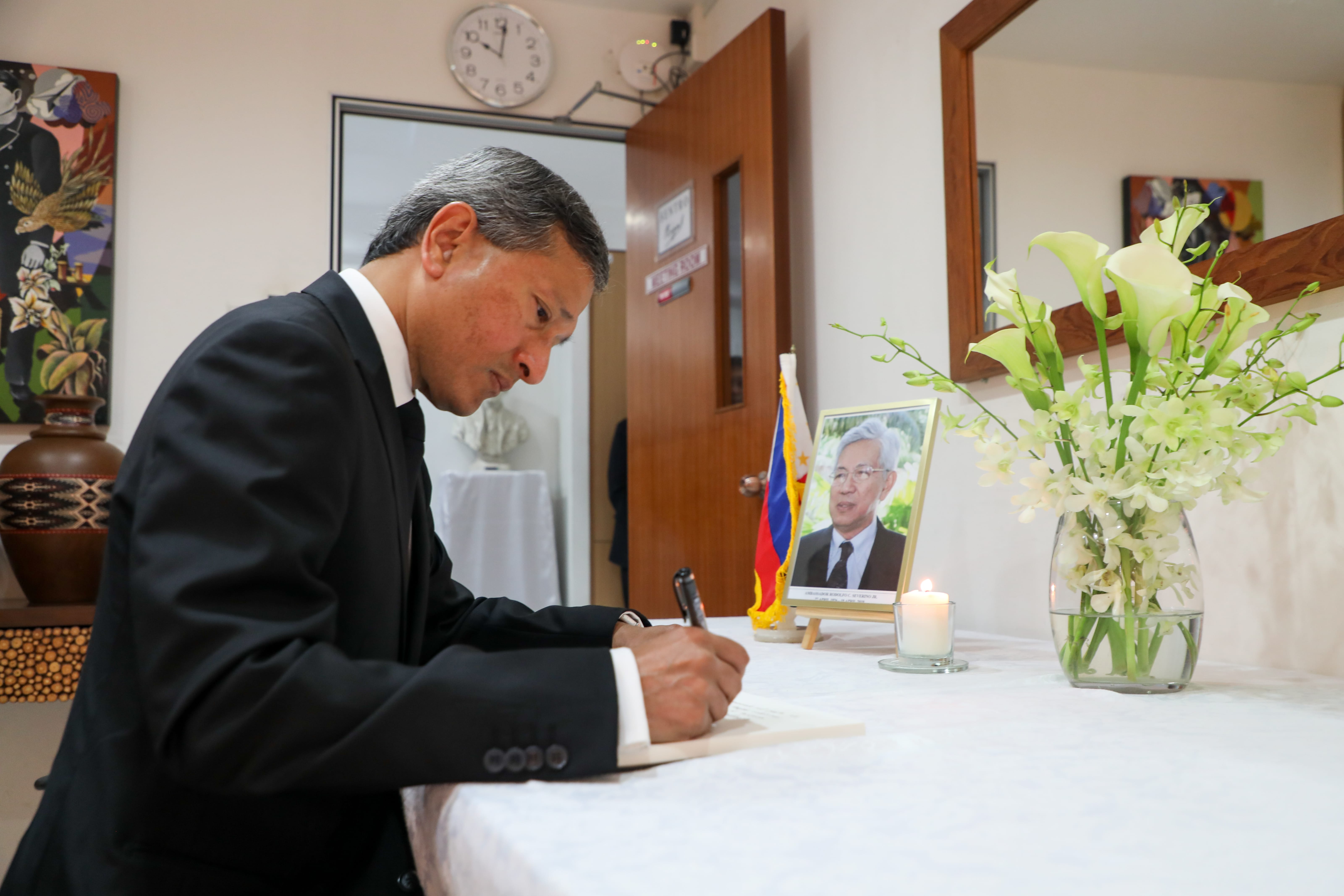 Man in suit writing; portrait with flag backdrop and flowers visible on a white table.